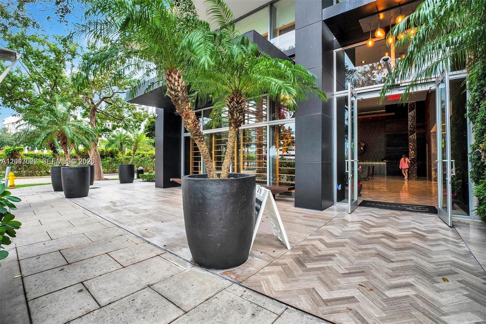 The exterior entrance of a modern building features large potted palm trees and herringbone flooring — 3470 E Coast Ave # H0904, Miami FL 33137, MIAMI, FL