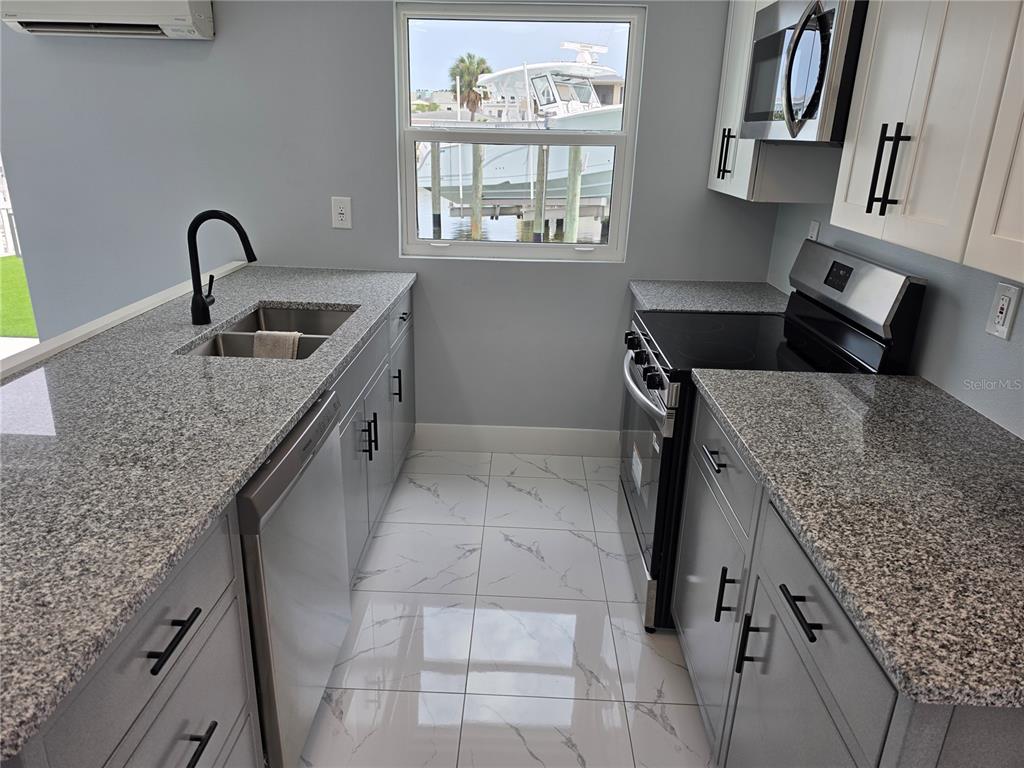 A galley kitchen features gray cabinets, granite countertops, stainless steel appliances, and a window view — 145 131 AVENUE E UNIT 2, MADEIRA BEACH, FL