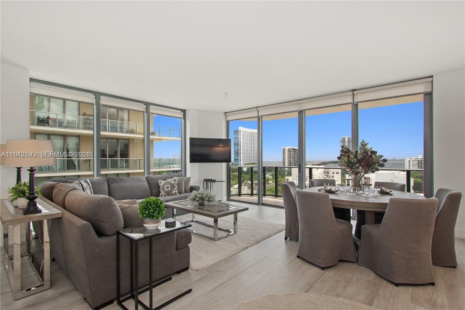 A modern living and dining area features floor-to-ceiling windows with a city and water view — 121 NE 34th St # 1008, Miami FL 33137, MIAMI, FL