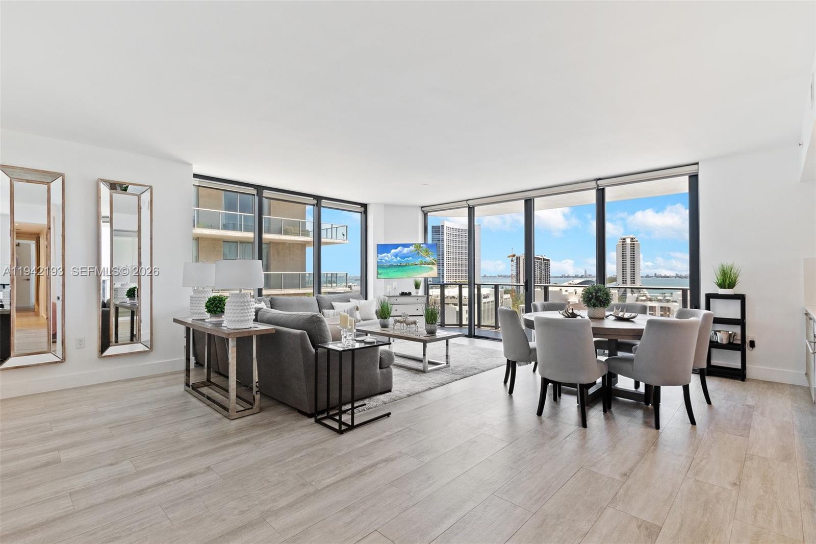 A modern living and dining room features floor-to-ceiling windows with expansive city and water views — 121 NE 34th St # 1008, Miami FL 33137, MIAMI, FL
