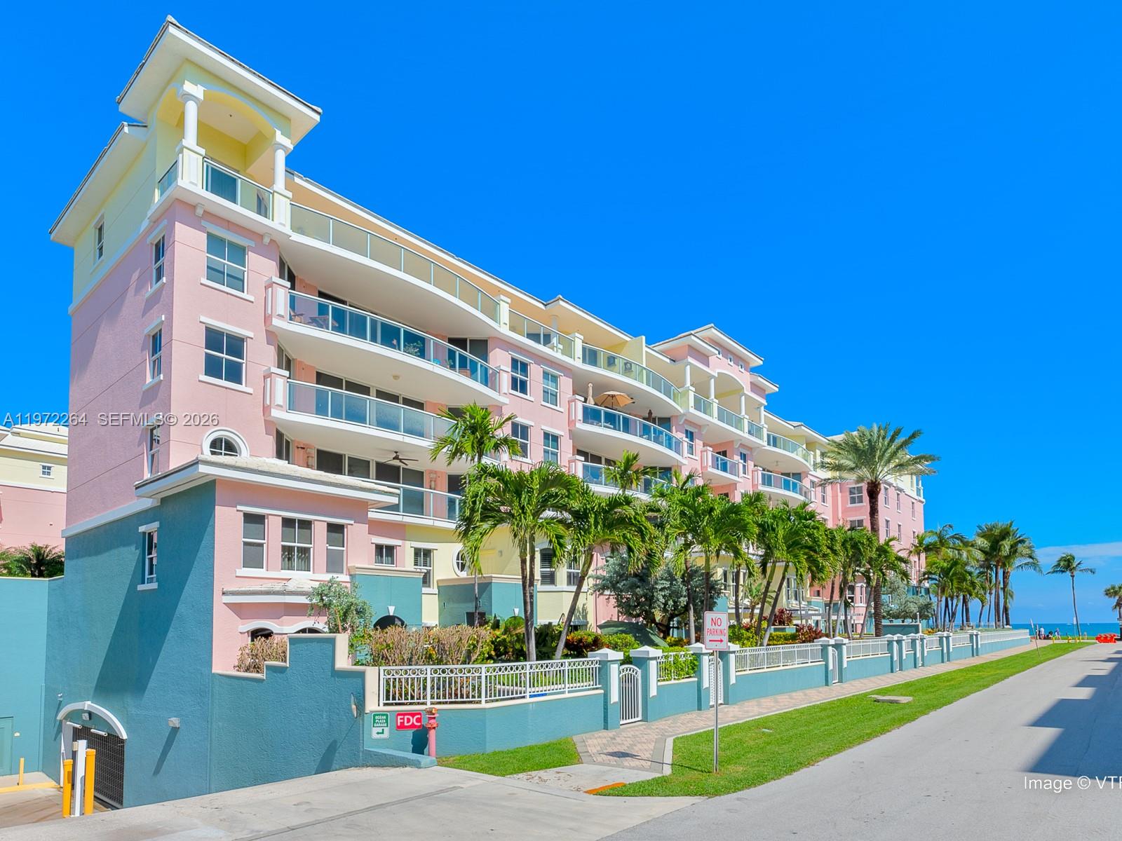 A multi-story pink and blue apartment building stands beside a paved road and palm trees — 2051 SE 3rd St, Deerfield Beach FL 33441, DEERFIELD BEACH, FL