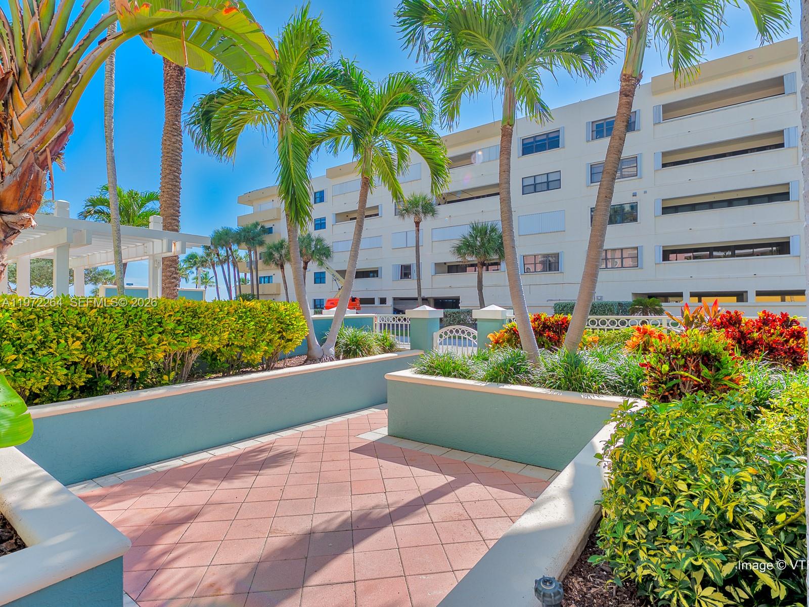 A paved courtyard features palm trees, landscaped planters, and a multi-story building in the background — 2051 SE 3rd St, Deerfield Beach FL 33441, DEERFIELD BEACH, FL
