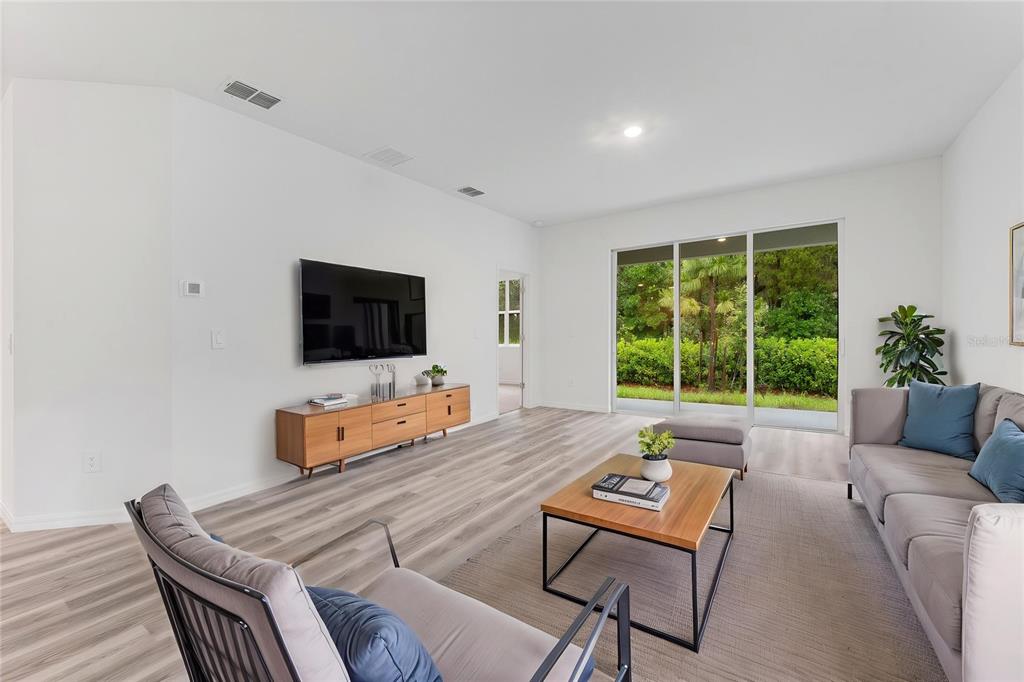 A bright living room features light wood-look flooring, a wall-mounted television, and sliding glass doors — 31351 PAPER BIRCH STREET, WESLEY CHAPEL, FL