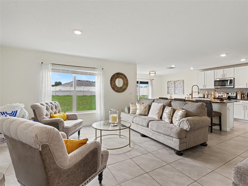 An open-concept living room features tile flooring, a gray sofa, two armchairs, and a kitchen — 16525 MOSAIC OAR DRIVE, WIMAUMA, FL