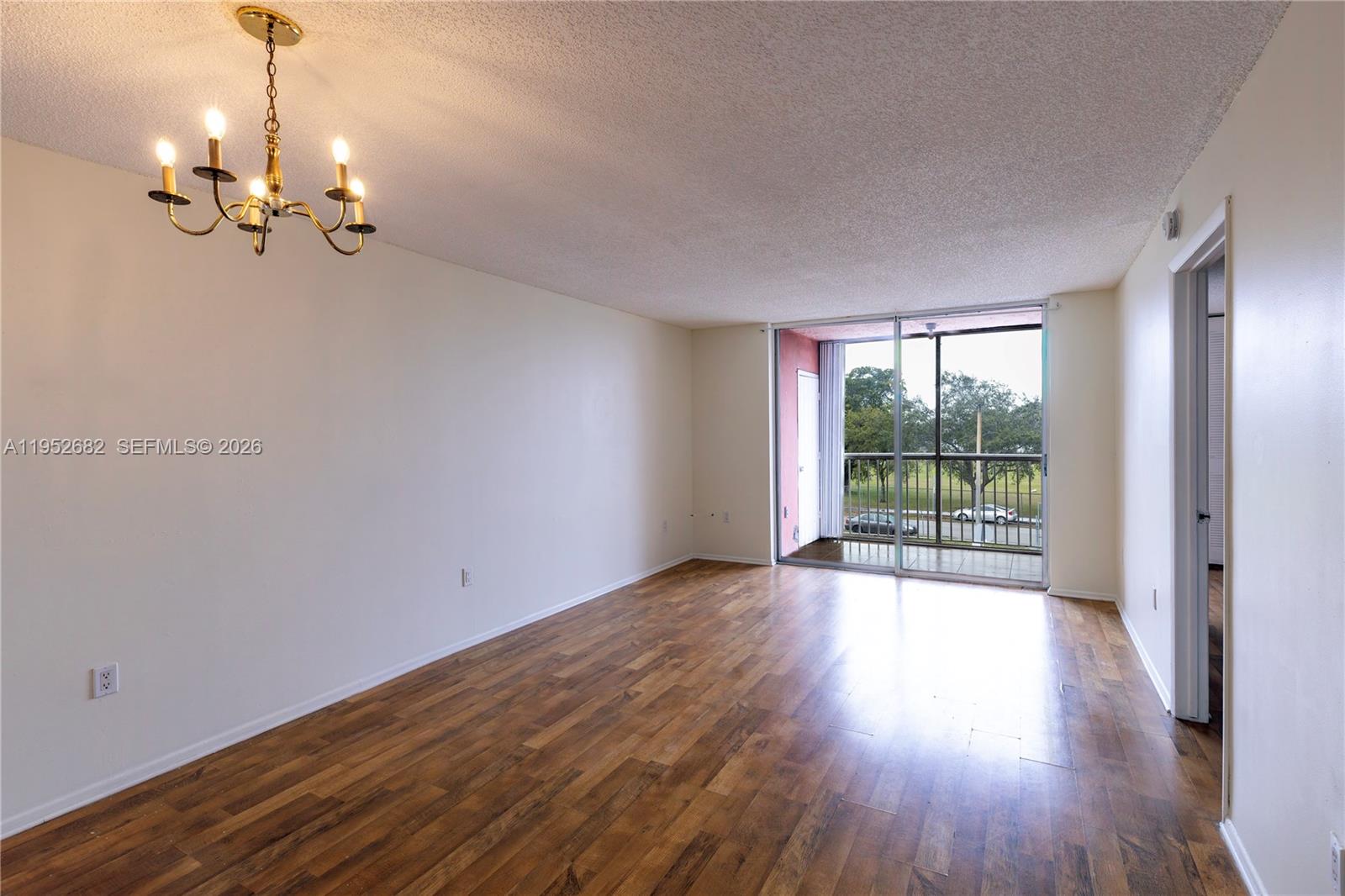 An empty living room features wood-look flooring, a brass chandelier, and a sliding glass balcony door — 19701 SW 114th Ave # 364, Miami FL 33157, MIAMI, FL