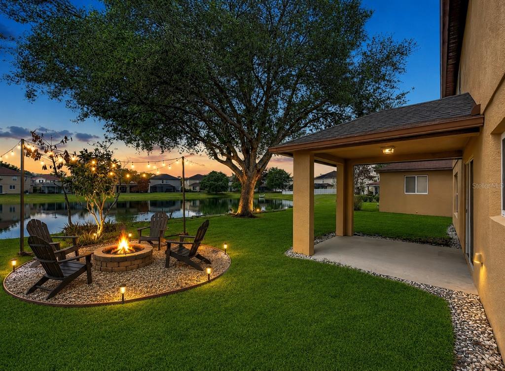 A backyard features a fire pit with chairs, string lights, a large tree, and pond — 14932 PERDIDO DRIVE, ORLANDO, FL