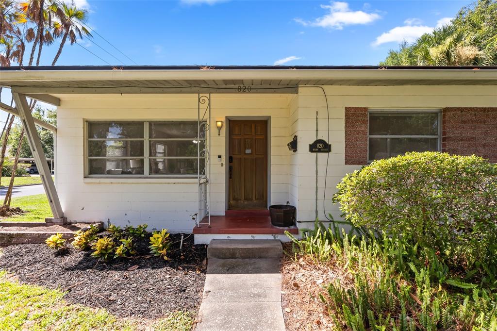The exterior of a single-story house features white siding, a wooden front door, and landscaping — 820 RUGBY STREET, ORLANDO, FL