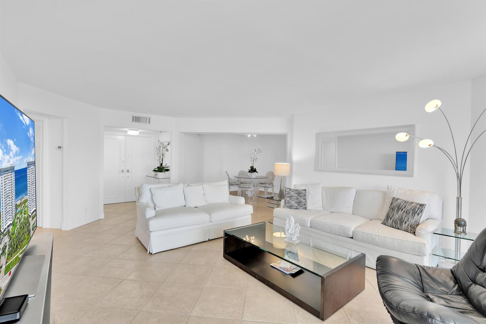A bright living room features white sofas, a glass coffee table, and tile flooring — 550 S Ocean Boulevard # 1003, Boca Raton FL 33432, BOCA RATON, FL