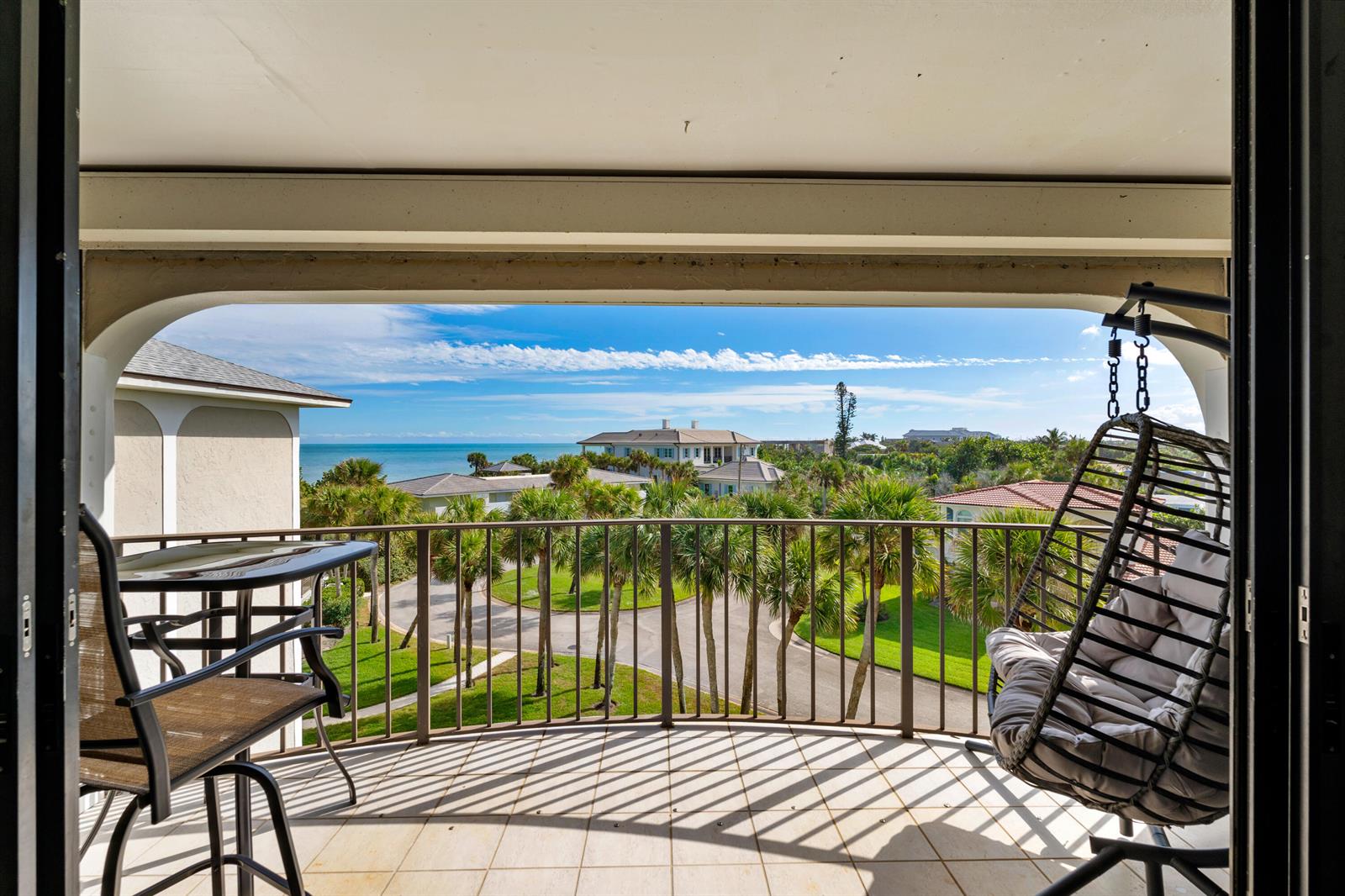 A balcony with a table, chairs, and a hanging swing chair overlooking palm trees and ocean — 5300 Highway A1a # 408, Vero Beach FL 32963, VERO BEACH, FL