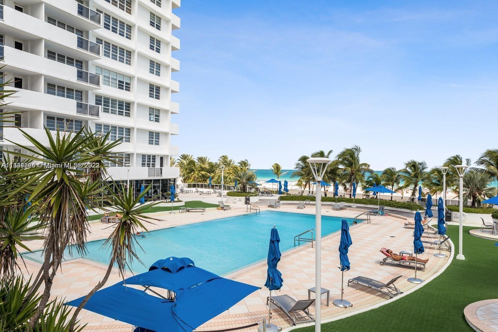A swimming pool area with lounge chairs and blue umbrellas sits beside a tall building — 100 Lincoln Rd # 1612, Miami Beach FL 33139, MIAMI BEACH, FL