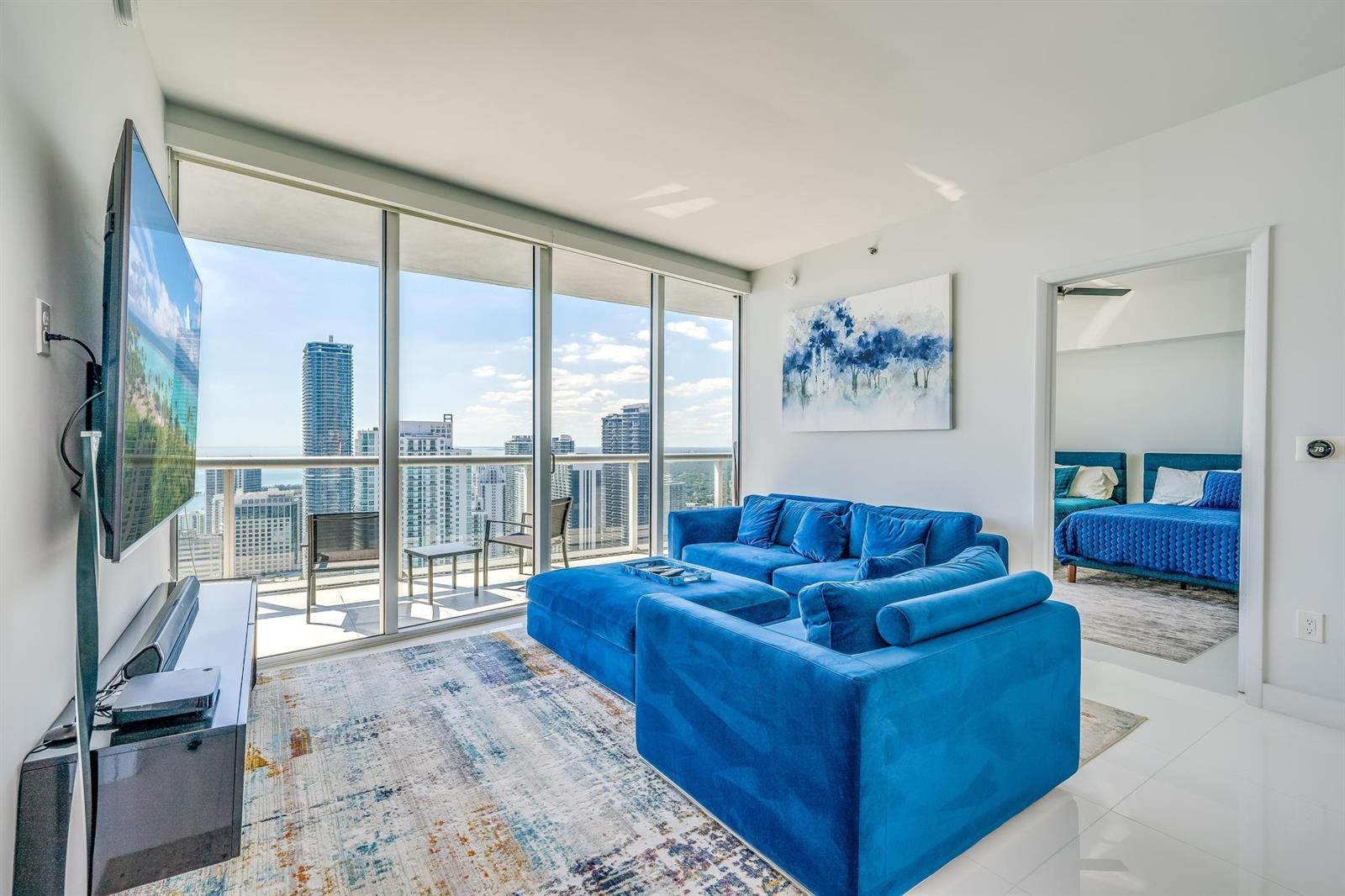 A living room with a blue sectional sofa, area rug, and floor-to-ceiling windows overlooking cityscapes — 495 Brickell Avenue # 5605, Miami FL 33131, MIAMI, FL