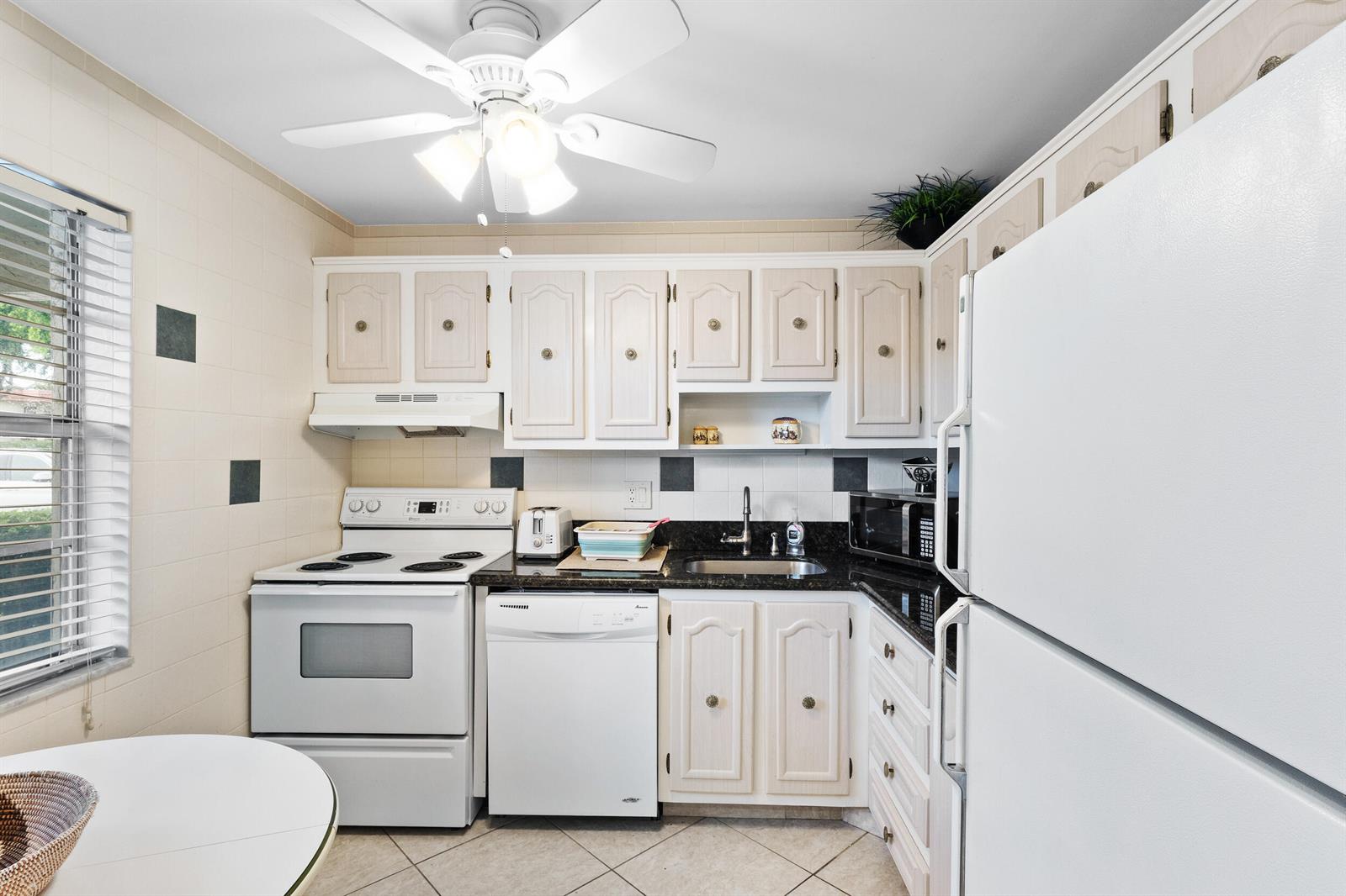 A kitchen features white cabinets, black countertops, white appliances, and a ceiling fan — 3 Abbey Lane # 102, Delray Beach FL 33446, DELRAY BEACH, FL