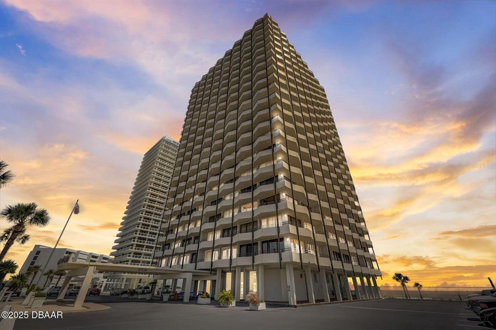 A tall, multi-story residential building with balconies stands against a sunset sky — 2900 N ATLANTIC AVENUE UNIT 1501, DAYTONA BEACH, FL