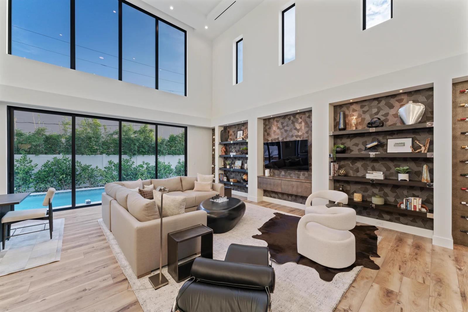 A modern living room features high ceilings, floor-to-ceiling windows, built-in shelving, and a pool view — 214 28th Street, West Palm Beach FL 33407, WEST PALM BEACH, FL