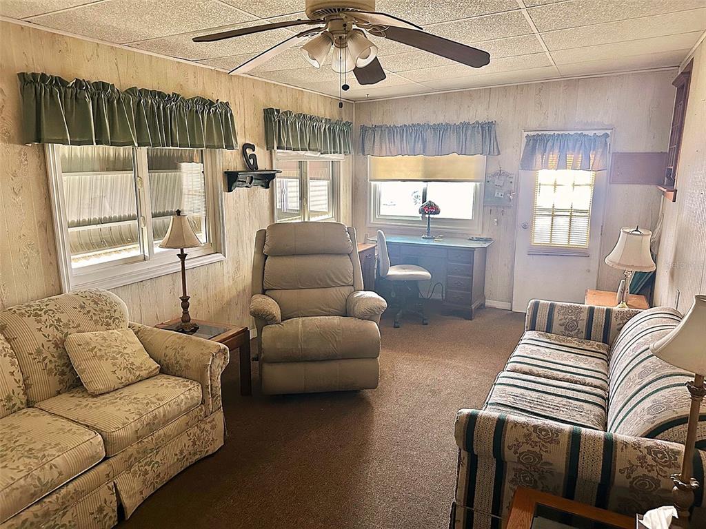 A living room with wood-paneled walls, carpeted flooring, a ceiling fan, and traditional furniture — 9790 66TH STREET N UNIT 265, PINELLAS PARK, FL