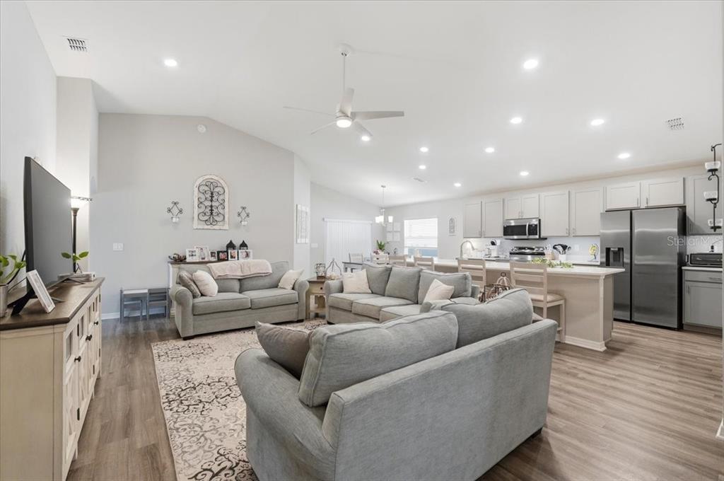An open-concept living room features gray sofas, wood-look flooring, and a kitchen with stainless appliances — 2610 SAN MARCO WAY, WINTER HAVEN, FL