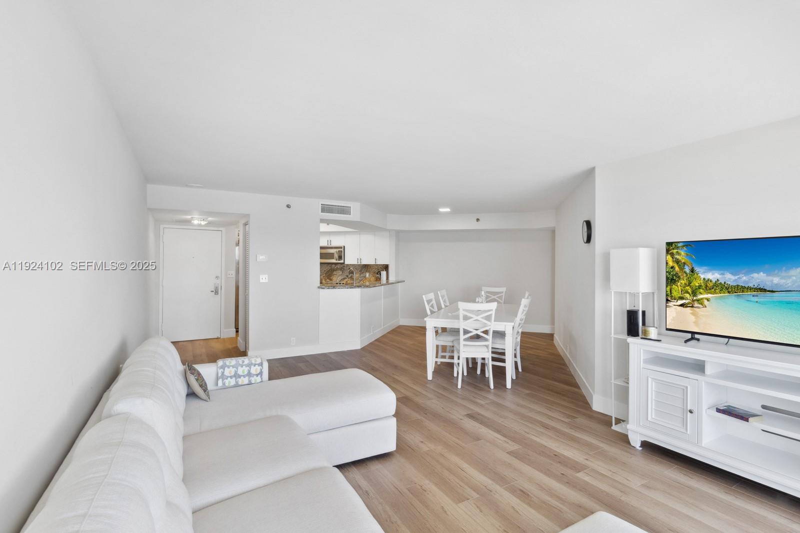 This open-concept living and dining area features white furniture, light wood flooring, and a kitchen — 6767 Collins Ave # 909, Miami Beach FL 33141, MIAMI BEACH, FL