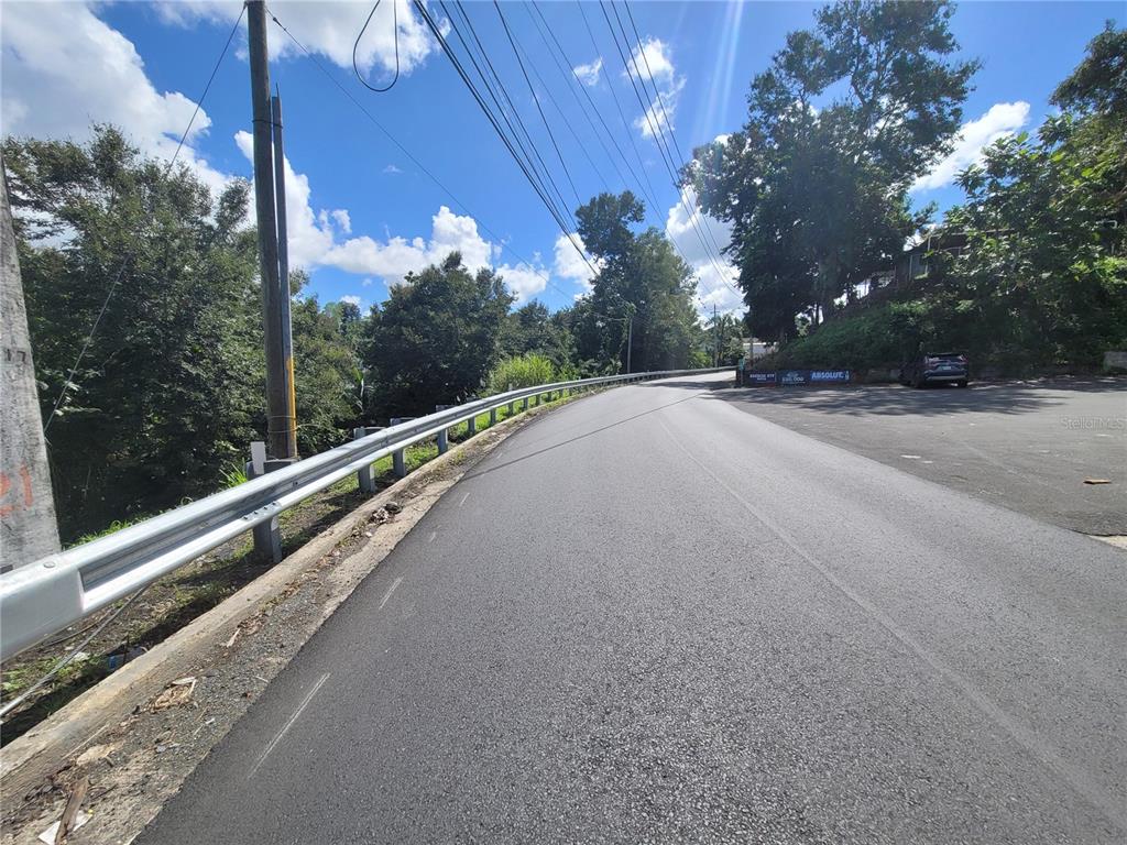 A paved road curves past a metal guardrail with trees and a parked car nearby — CARR 829, KM 2.3 BO. BUENA VISTA, BAYAMON, PR
