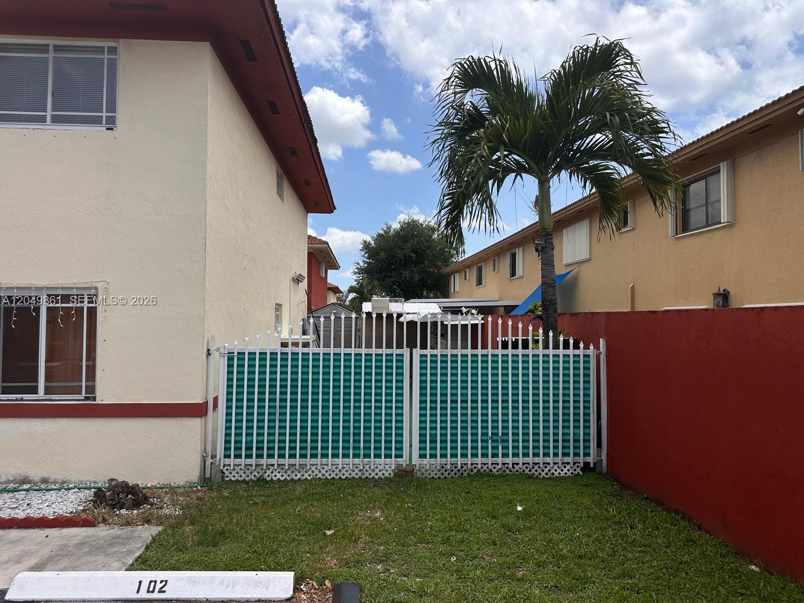 A white metal gate with green privacy slats stands between two residential buildings — 11053 W Okeechobee Rd # 102, Hialeah Gardens FL 33018, HIALEAH GARDENS, FL