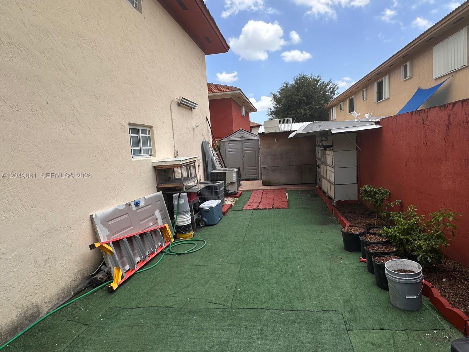 A narrow outdoor side yard features artificial turf, a red wall, and various storage items — 11053 W Okeechobee Rd # 102, Hialeah Gardens FL 33018, HIALEAH GARDENS, FL