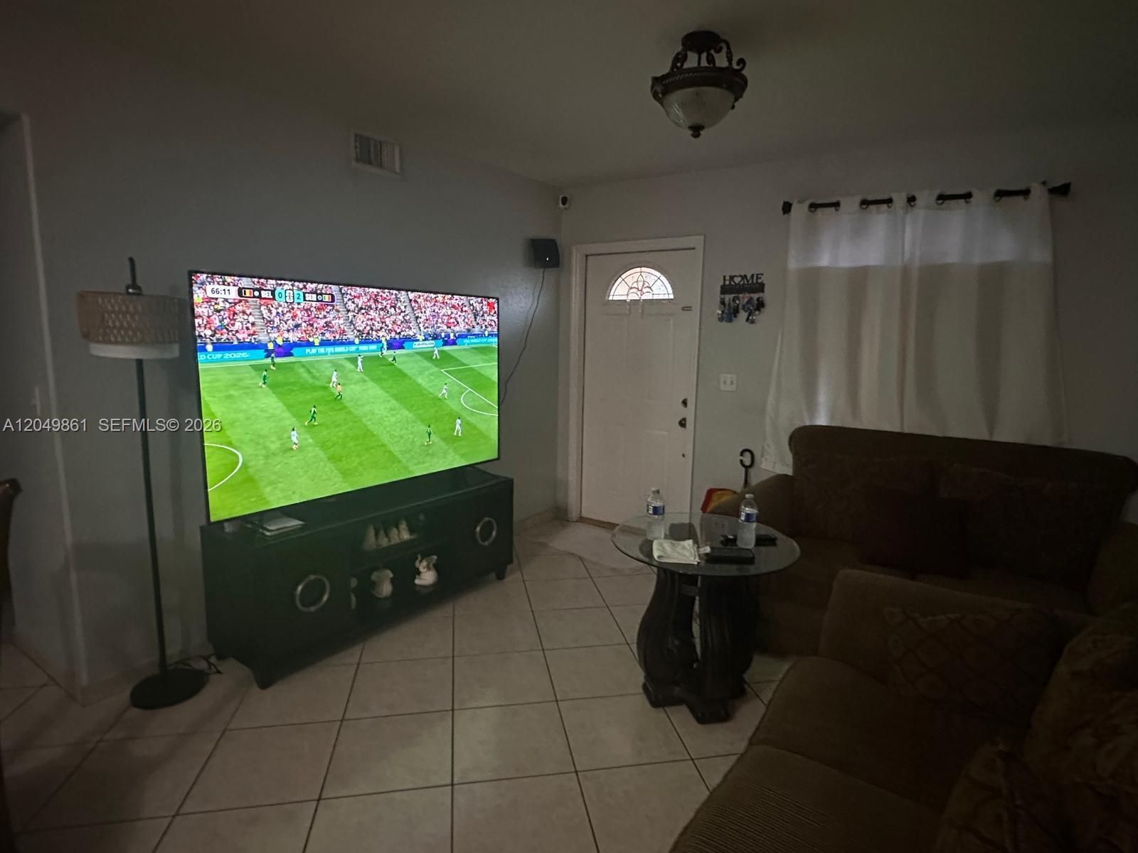 A living room features a large television on a dark console, tiled floors, and a sofa — 11053 W Okeechobee Rd # 102, Hialeah Gardens FL 33018, HIALEAH GARDENS, FL