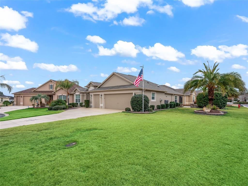 A tan single-story house with a three-car garage and an American flag in the yard — 3556 MISTLETOE COURT, THE VILLAGES, FL