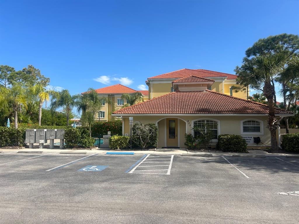 A tan two-story building with a red tile roof stands behind a paved parking lot — 940 COOPER STREET UNIT 302, VENICE, FL