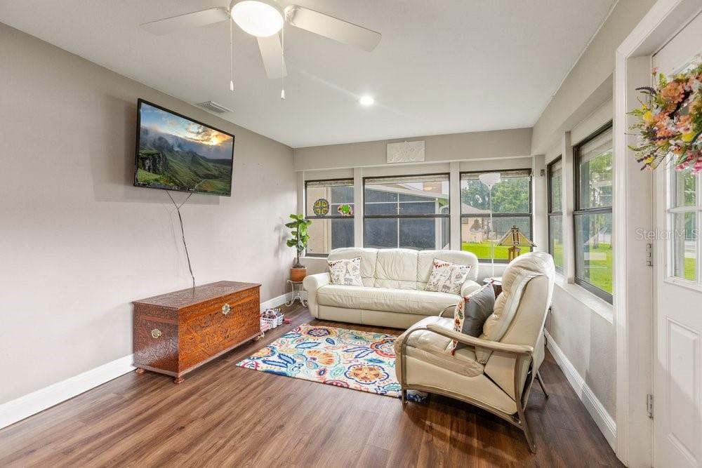 A sunroom features wood-look flooring, a white sofa, an armchair, and a wall-mounted television — 13502 WHITBY ROAD, HUDSON, FL