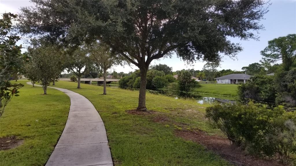 A paved walkway winds through a grassy park area alongside a canal and mature trees — 1143073705 NEW MILFORD STREET, NORTH PORT, FL