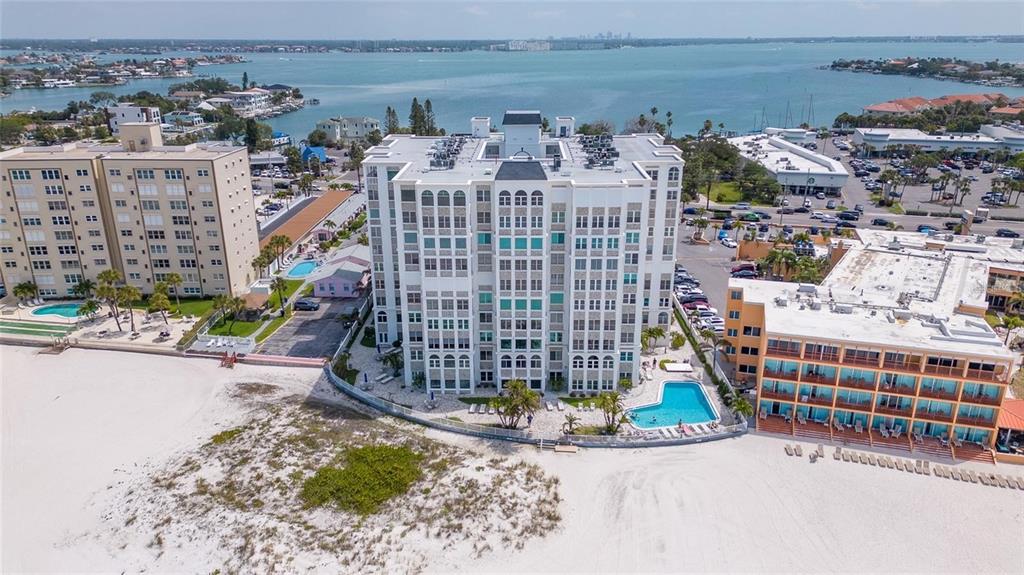 An aerial view shows a multi-story white apartment building situated between a beach and a parking lot — 4950 GULF BOULEVARD UNIT 501, ST PETE BEACH, FL