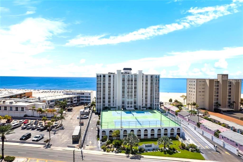 An aerial view shows a tall beachfront condominium building with tennis courts and parking — 4950 GULF BOULEVARD UNIT 501, ST PETE BEACH, FL