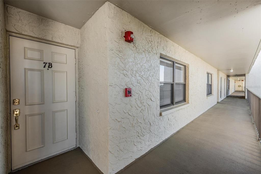 An exterior view of an apartment building hallway featuring a white door labeled 7B — 2900 COVE CAY DRIVE UNIT 7B, CLEARWATER, FL