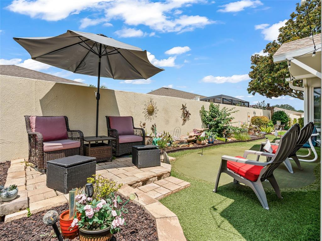 A backyard patio features a stone seating area with an umbrella, chairs, and putting green — 17770 SE 85TH ELLERBE AVENUE, THE VILLAGES, FL
