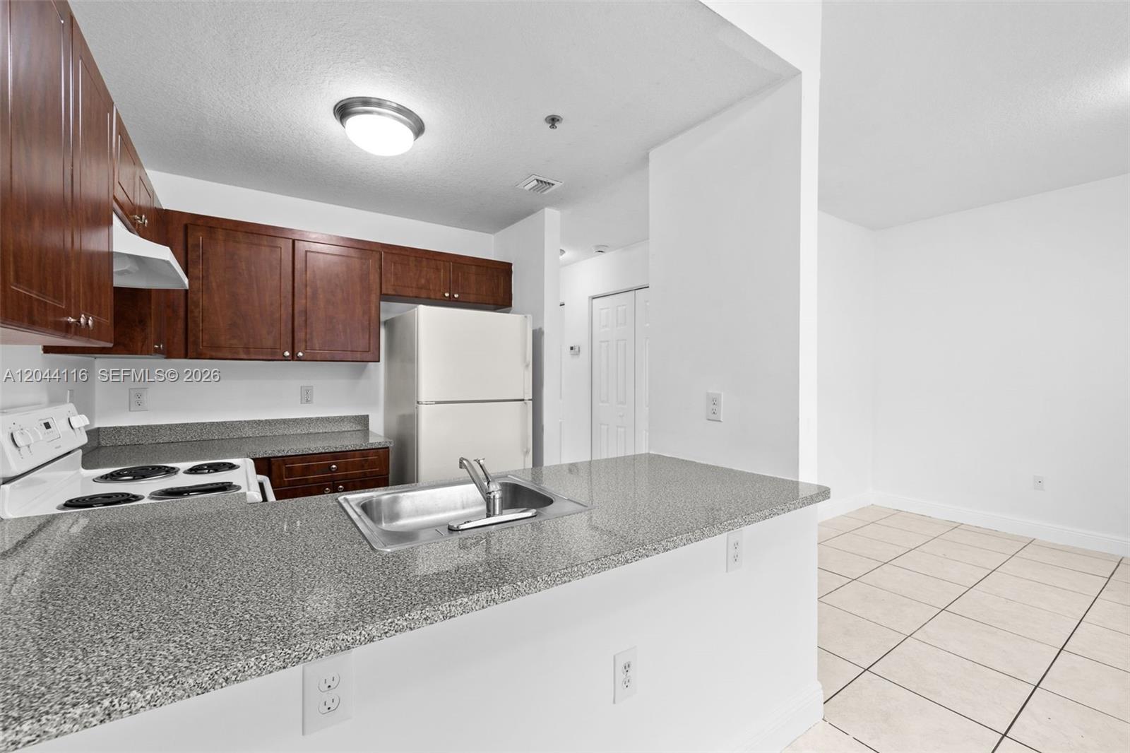 A kitchen features dark wood cabinets, granite countertops, a white refrigerator, and tiled flooring — 2421 W 78th St # 104, Hialeah FL 33016, HIALEAH, FL