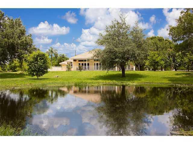 A tan house with a pool enclosure sits behind a pond surrounded by green trees — 2088 Appaloosa Trail, Wellington FL, WELLINGTON, FL