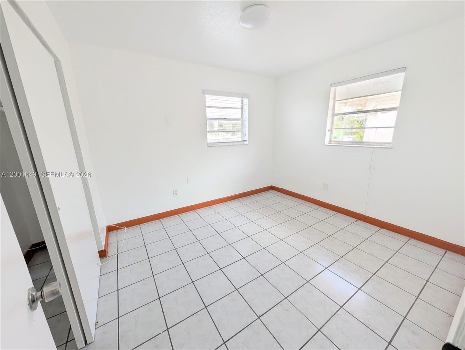 An empty room features white walls, tiled flooring, and orange baseboards with two windows — 1551 NE 8th St # 8, Homestead FL 33033, HOMESTEAD, FL
