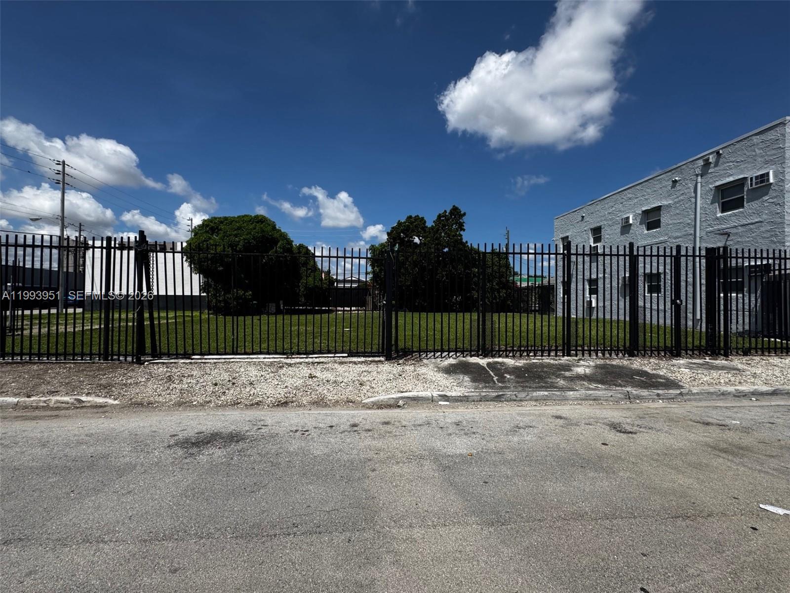 A fenced, vacant grassy lot sits next to a multi-story gray building under a blue sky — 2410 NW 7th Ct, Miami FL 33127, MIAMI, FL