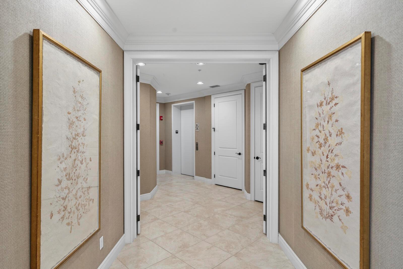 A hallway with beige textured wallpaper, framed floral art, and tile flooring leads to elevators — VERO BEACH, FL