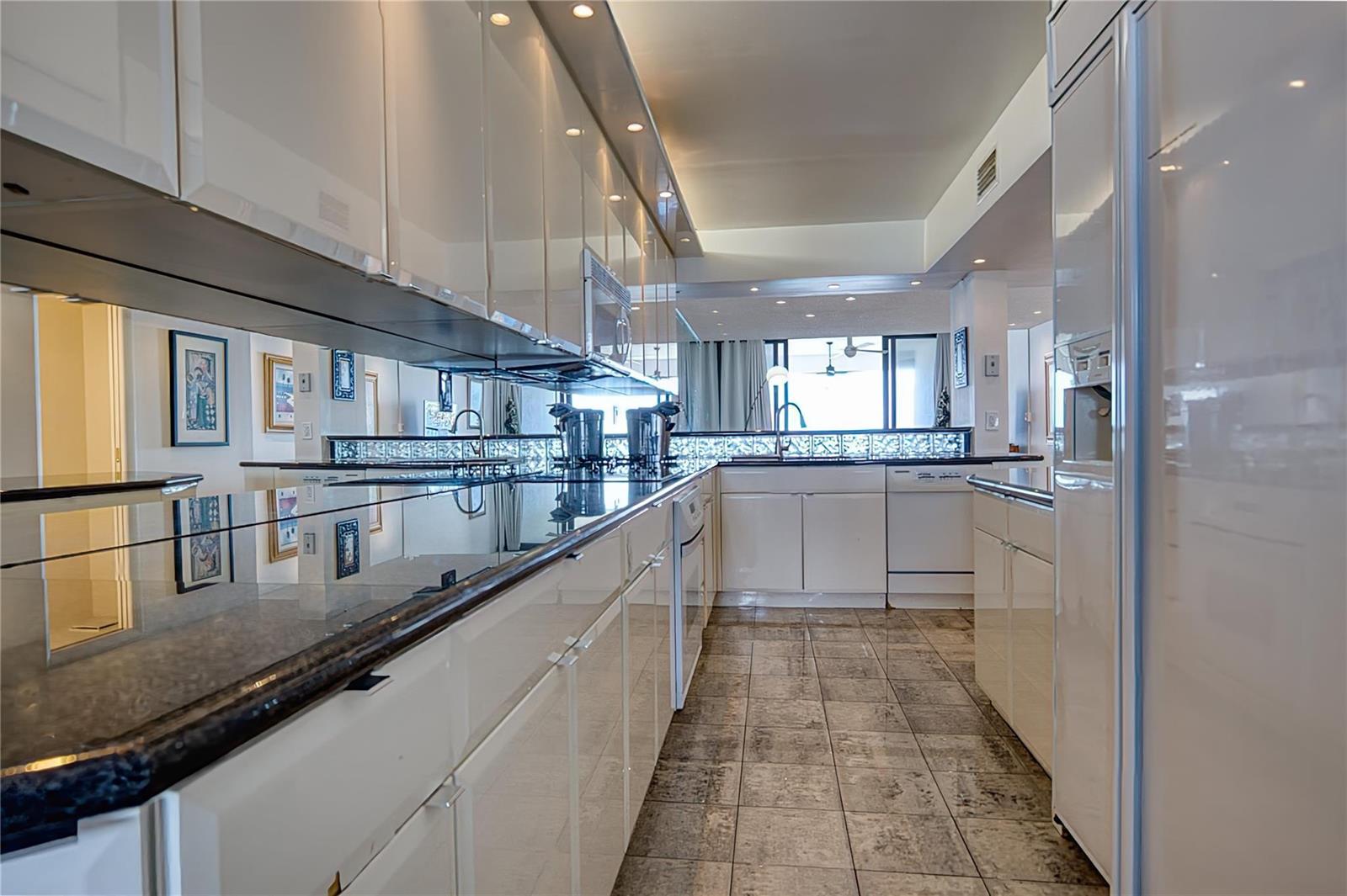 A galley kitchen features white high-gloss cabinets, dark countertops, and tiled flooring — 520 N Ocean Boulevard # 13, Pompano Beach FL 33062, POMPANO BEACH, FL