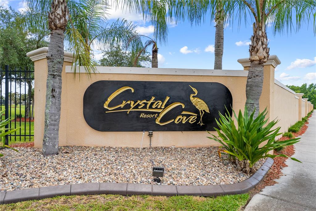 A tan stucco wall features a black sign with gold lettering reading Crystal Cove Resort — 4704 RUBY RED LANE, KISSIMMEE, FL