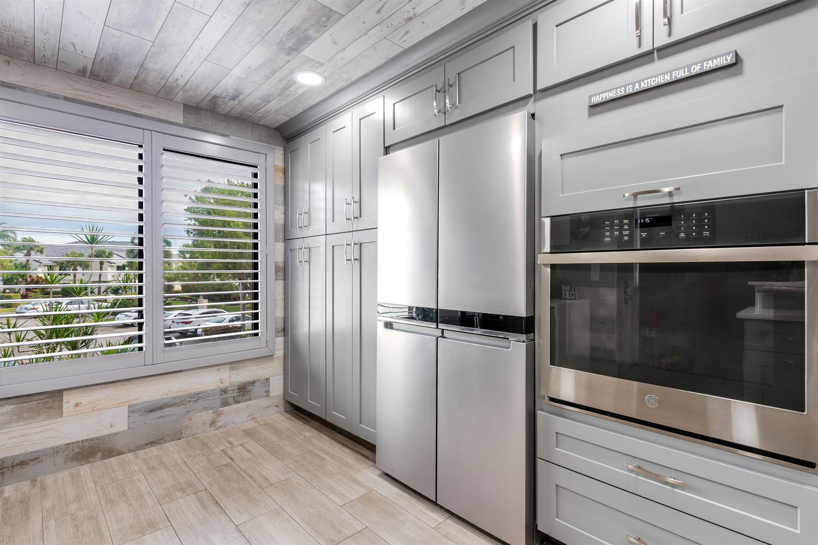 A kitchen features grey cabinetry, stainless steel appliances, wood-look tile flooring, and a window with shutters — 7418 Victory Lane # 9805, Delray Beach FL 33446, DELRAY BEACH, FL
