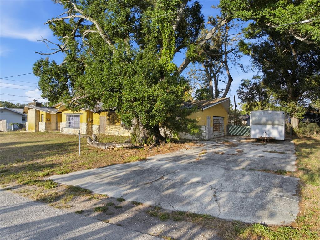 A yellow single-story house with a stone facade, a concrete driveway, and large mature trees — 8181 61ST LANE N, PINELLAS PARK, FL