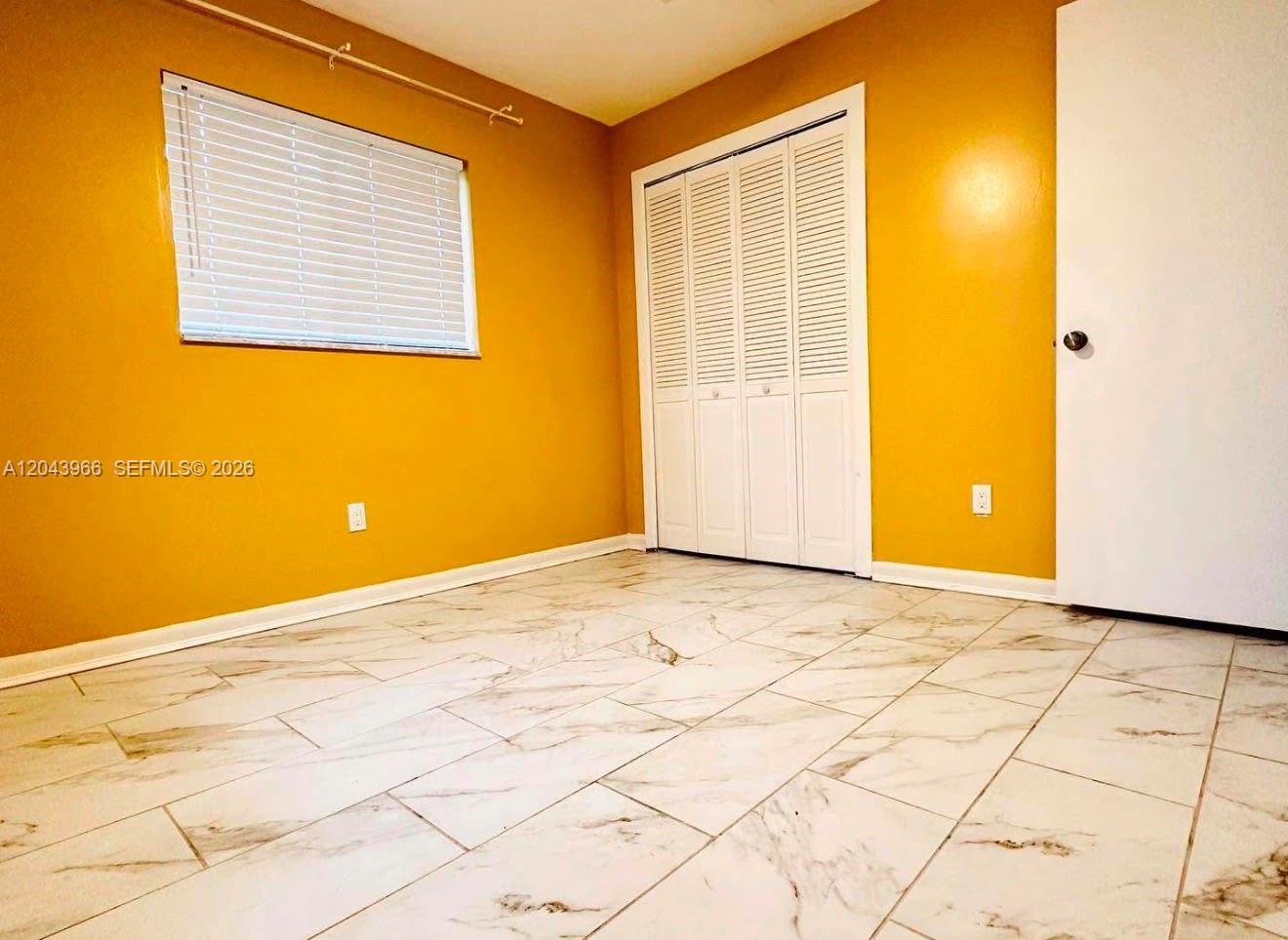 An empty room with yellow walls, white tile flooring, a window with blinds, and closets — 709 NW Cuxhaven St, Palm Bay FL 32907, PALM BAY, FL