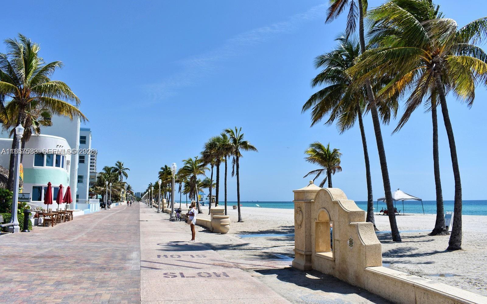 A paved boardwalk runs alongside a sandy beach lined with palm trees under a clear sky — 3801 S Ocean Dr # 12M, Hollywood FL 33019, HOLLYWOOD, FL
