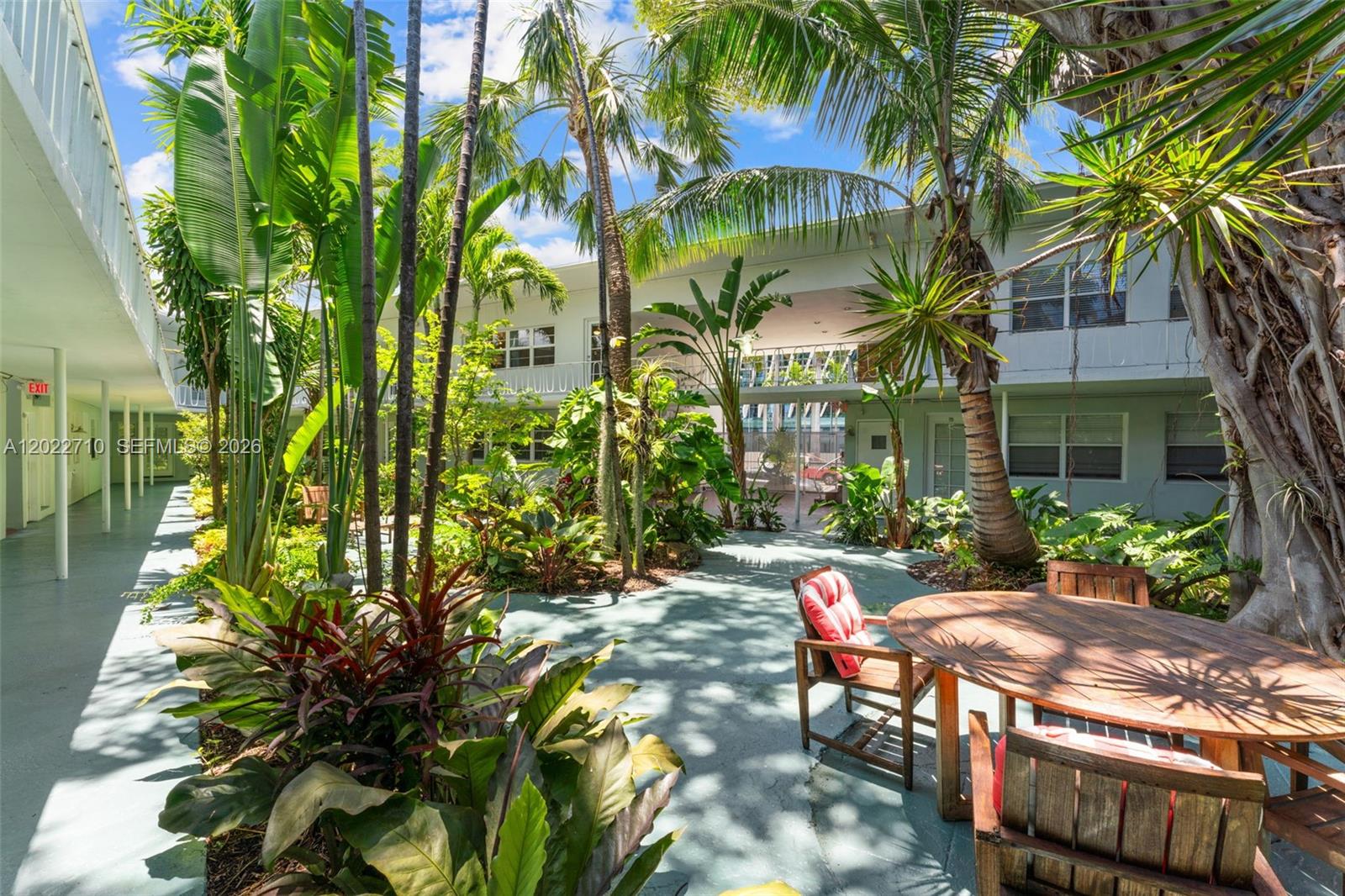 A lush, tropical courtyard features a wooden patio table and chairs surrounded by palm trees — 1925 Washington Ave # 16, Miami Beach FL 33139, MIAMI BEACH, FL