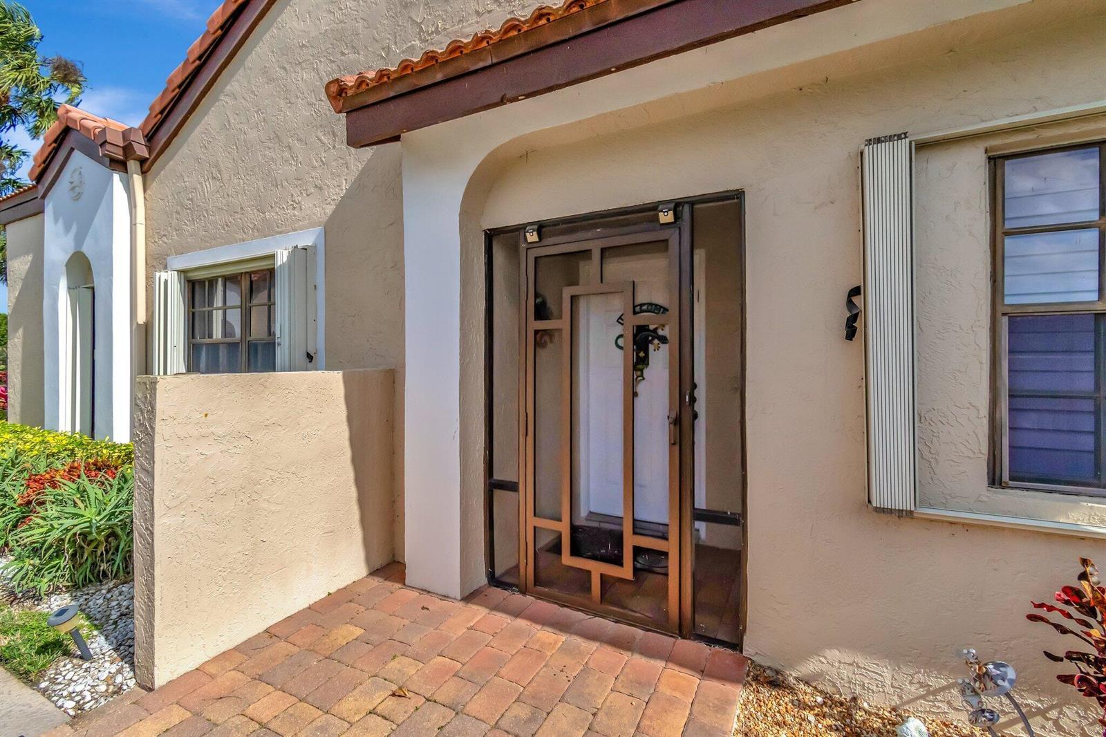 A tan stucco house exterior features a brick walkway, a decorative screen door, and shutters — 8240 Waterline Drive, Boynton Beach FL 33472, BOYNTON BEACH, FL