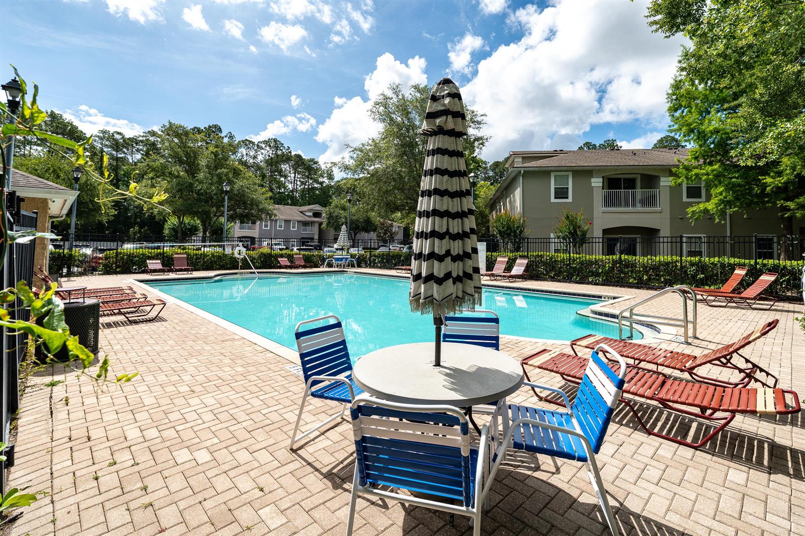 An outdoor swimming pool area features lounge chairs, a patio table with an umbrella, and buildings — 6069 Maggies Circle # 103, Jacksonville FL 32244, JACKSONVILLE, FL