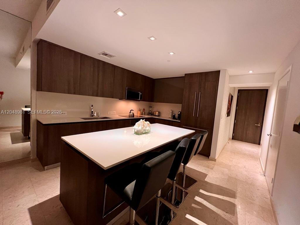 A modern kitchen features dark wood cabinetry, a white island, and light stone flooring — 88 SW 7th St # 808, Miami FL 33130, MIAMI, FL