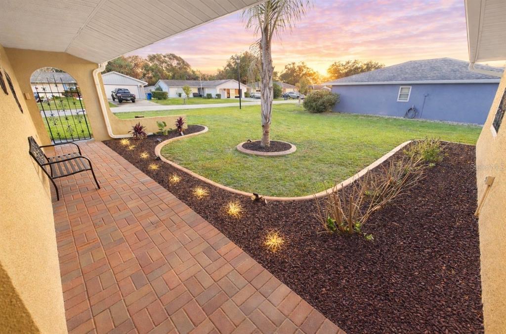 A paved patio overlooks a grassy yard with a palm tree and landscaped garden beds — 7051 LEXINGTON CIR, BROOKSVILLE, FL