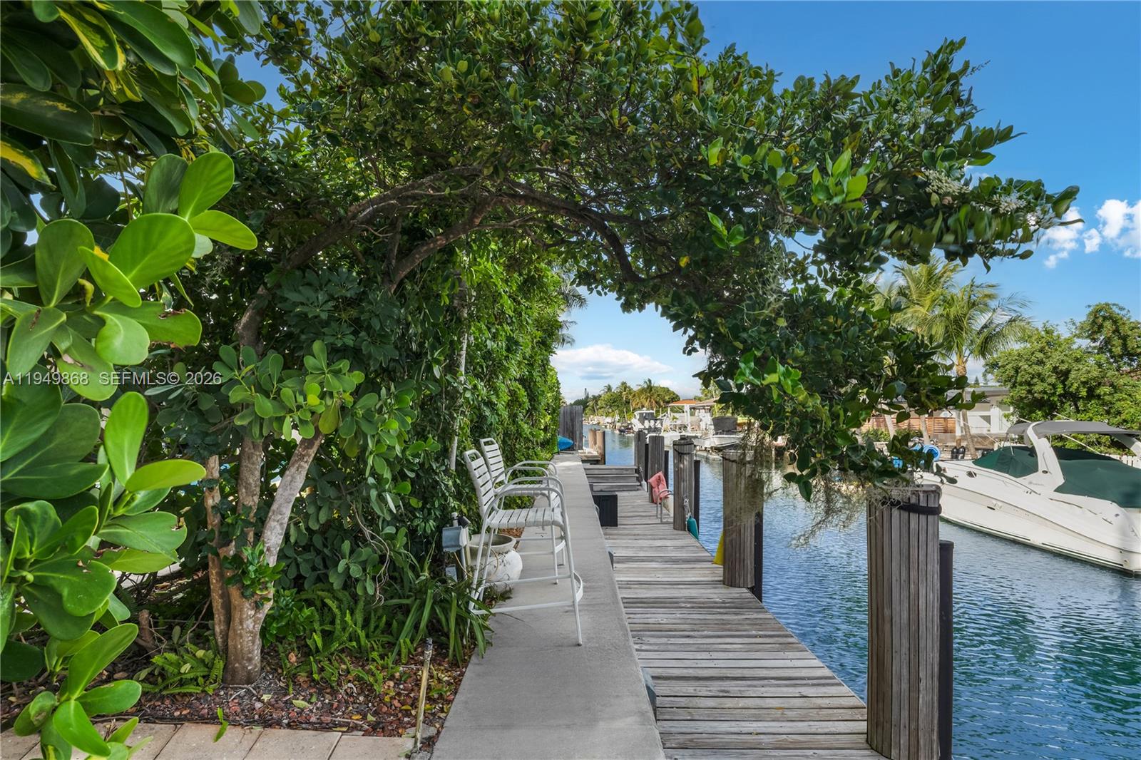 A wooden dock with white chairs sits beside a canal with a docked boat — 1155 S Biscayne Point Rd, Miami Beach FL 33141, MIAMI BEACH, FL